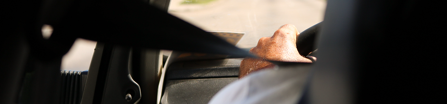 image of a person driving with their seatbelt on, focused on the driver's left hand, seatbelt and the windshield showing the road ahead.